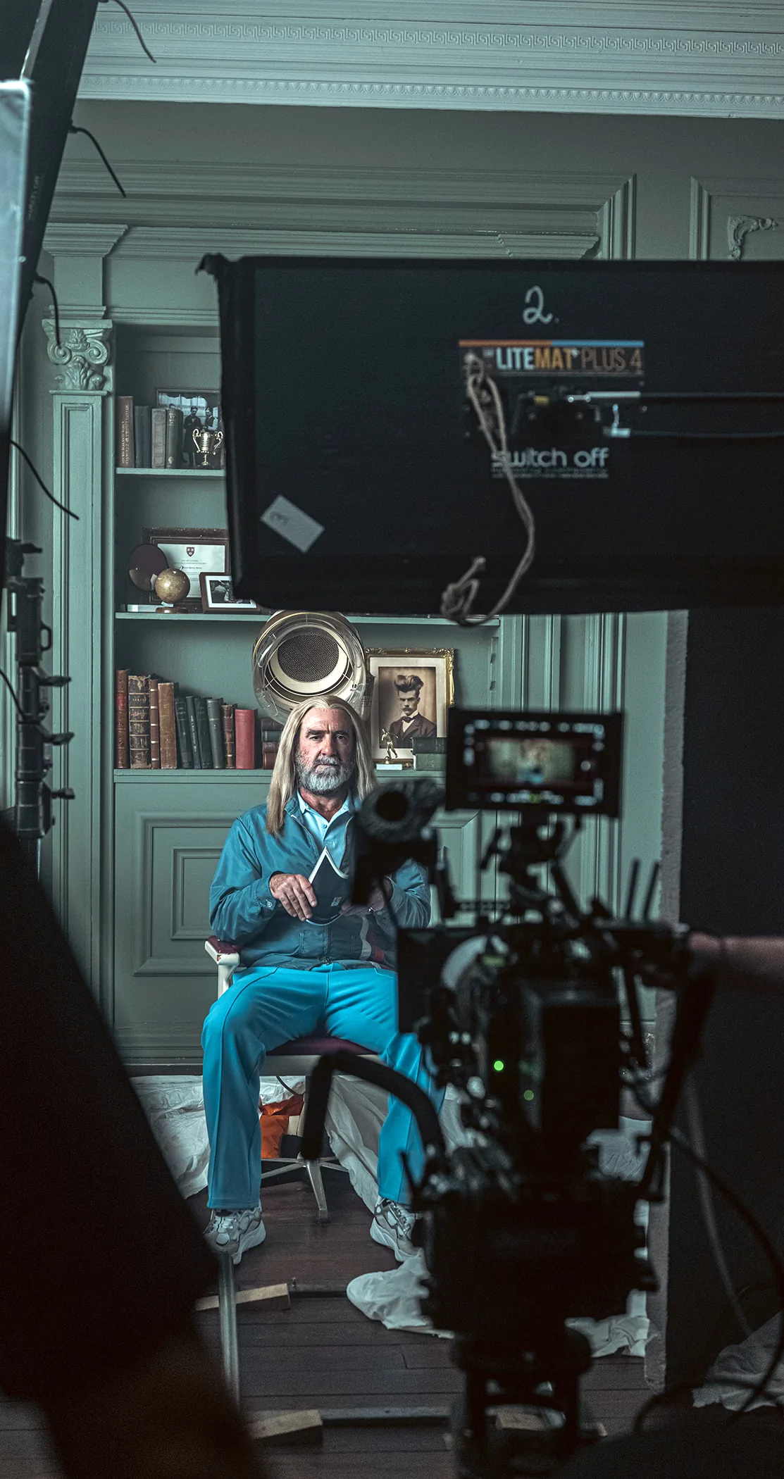 A man with long grey hair seated in a panelled room behind a cinema camera