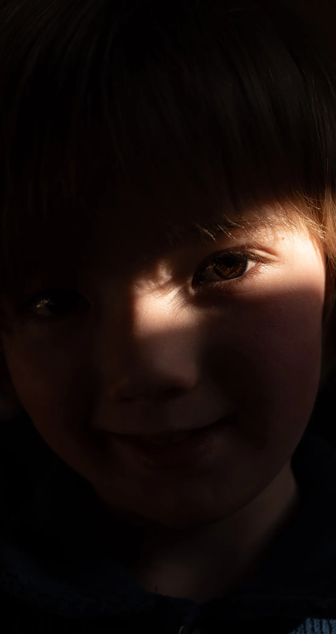 A smiling boy, his face half lit against deep shadow