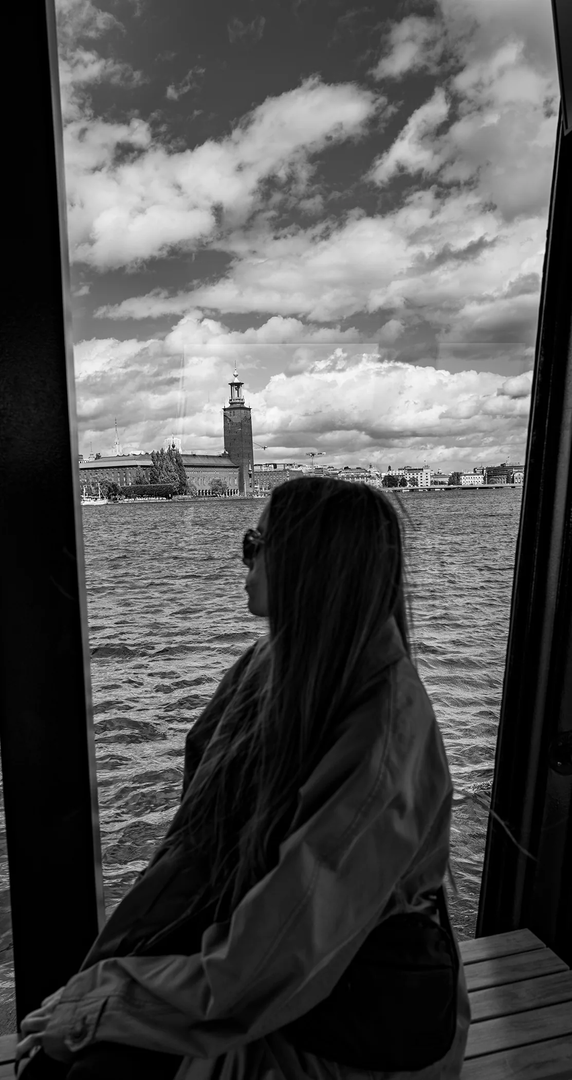 A woman in sunglasses looking across the water to Stockholm City Hall, in black and white