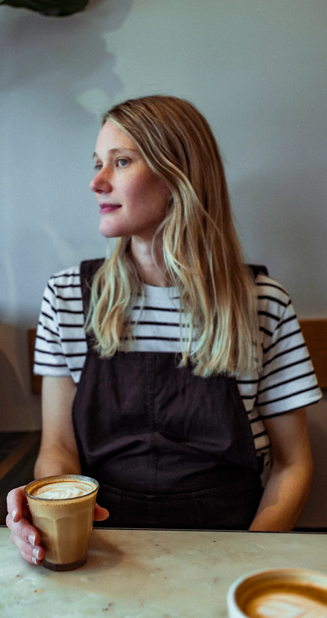 A woman in a striped top and apron holding a coffee, looking out of frame